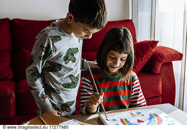 Smiling sister painting rainbow by brother in living room at home