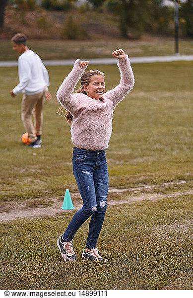 Smiling sister with arms raised standing on grassy field while brother playing soccer in background at park