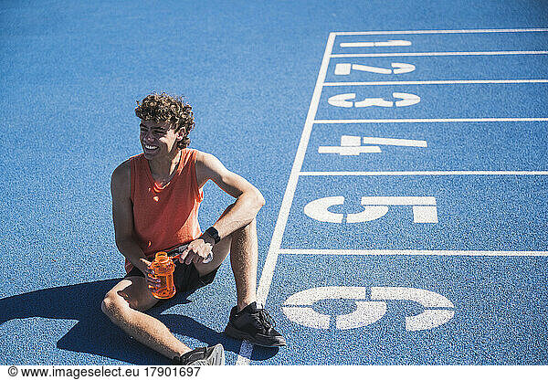 Smiling sportsman holding water bottle sitting by starting line on track