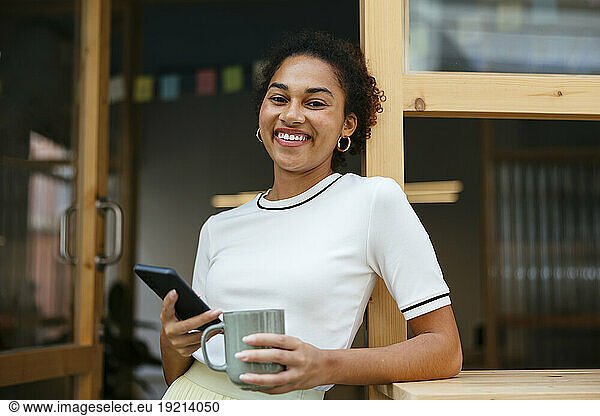 Smiling student standing with coffee cup and smart phone in doorway