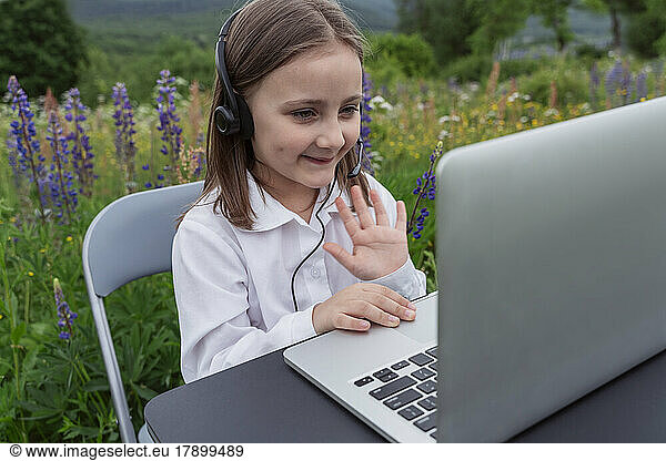 Smiling student waving on video call through laptop in meadow