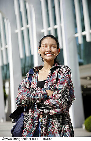 Smiling student with arms crossed standing in front of university