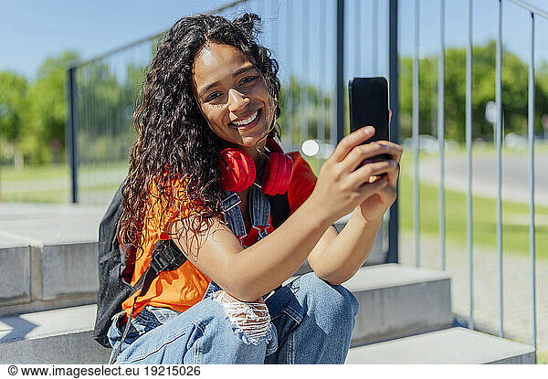 Smiling student with curly hair holding smart phone on steps in campus