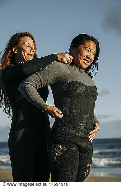 Smiling surfer helping woman putting on wetsuit at beach