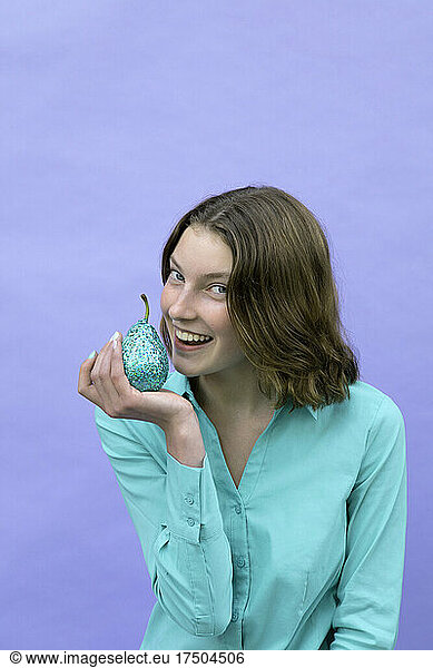 Smiling teenage girl holding pear in front of mouth against lavender background