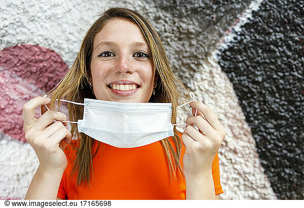 Smiling teenage girl holding protective face mask against graffiti wall
