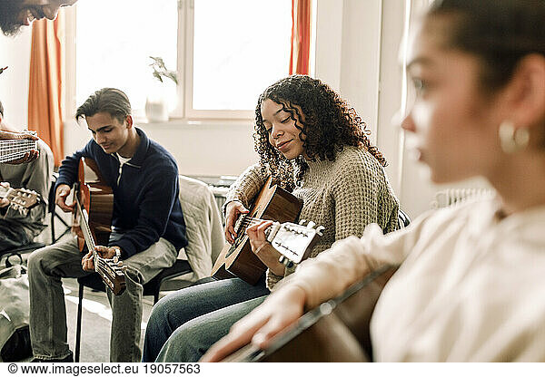 Smiling teenage girl practicing guitar with friends in music class at high school