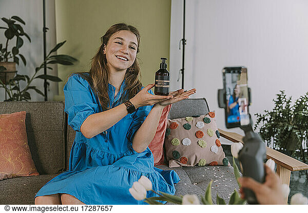 Smiling teenage girl presenting beauty product while vlogging at home