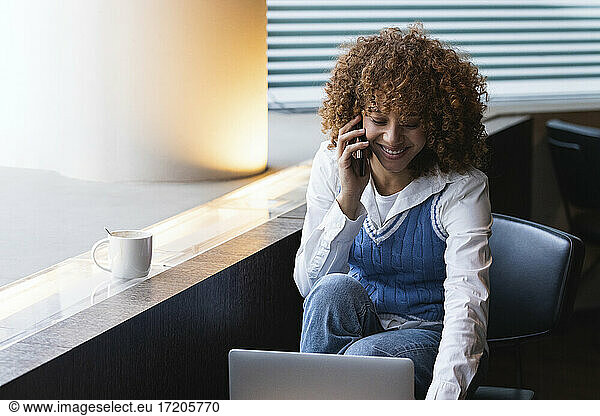 Smiling teenage girl talking on mobile phone while using laptop at cafe