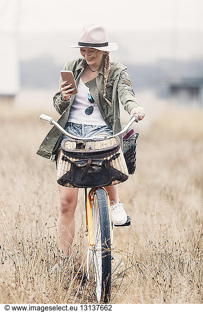 Smiling teenage girl using smart phone while sitting on bicycle amidst grassy field