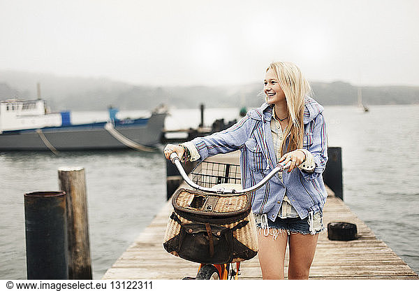 Smiling teenage girl with bicycle walking on pier over lake