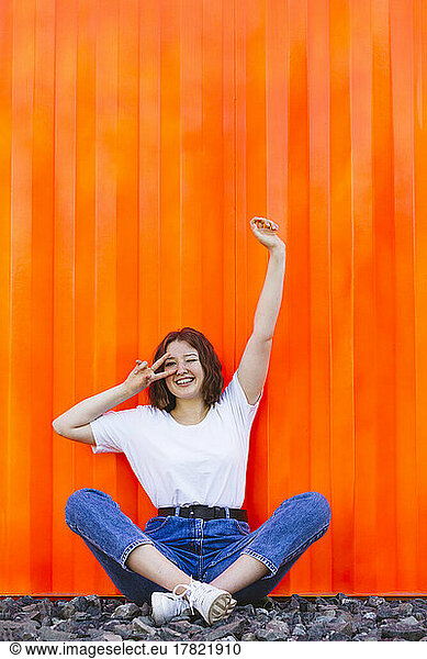 Smiling teenage girl with hand raised gesturing peace sign sitting in front of orange cargo container