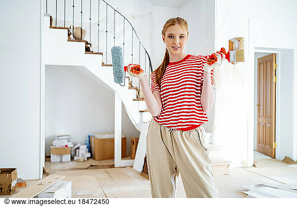 Smiling teenage girl with paint roller and brushes standing at home