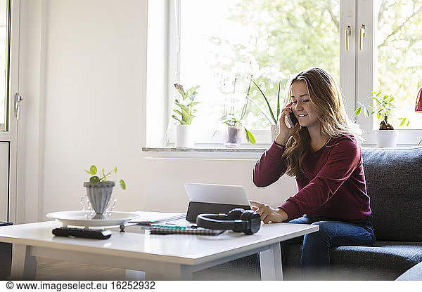 Smiling teenager talking on phone while using digital tablet at home