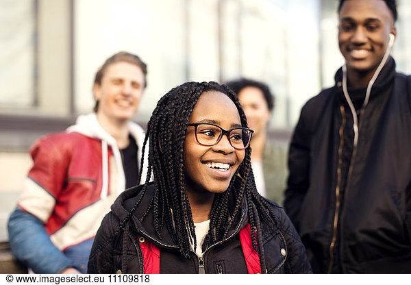 Smiling teenager with braided hair wearing eyeglasses standing against friends in city