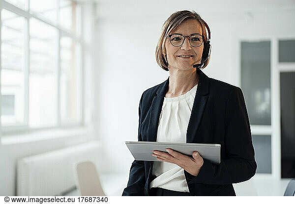 Smiling telecaller wearing headset holding tablet PC in office