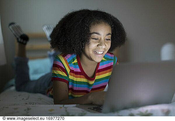 Smiling ten year-old bi-racial girl working on her apple laptop on bed