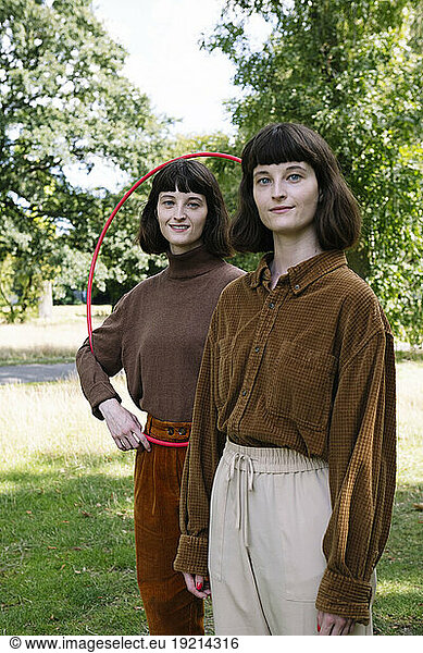 Smiling twin sisters standing with plastic hoop at park