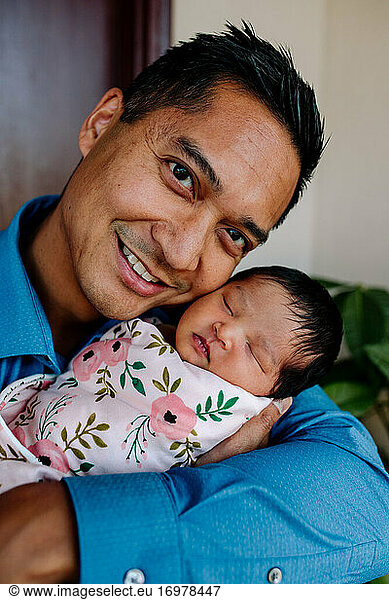 Smiling unshaven asian dad holds sleeping newborn daughter close