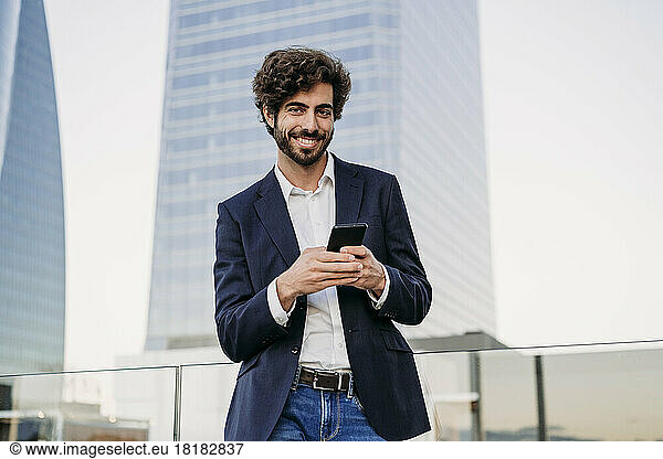 Smiling urban businessman with smart phone in front of building
