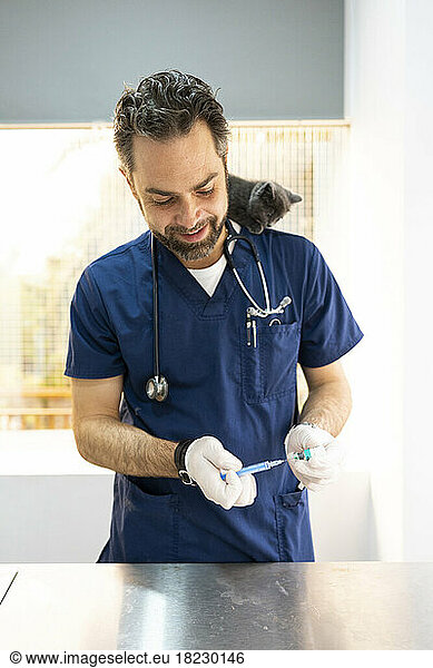 Smiling veterinarian preparing syringe with cat sitting on shoulder at clinic
