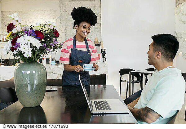 Smiling waitress serving coffee to customer sitting with laptop in cafe