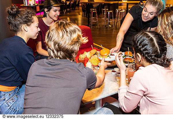 Smiling waitress serving food to multi-ethnic teenagers sitting at restaurant