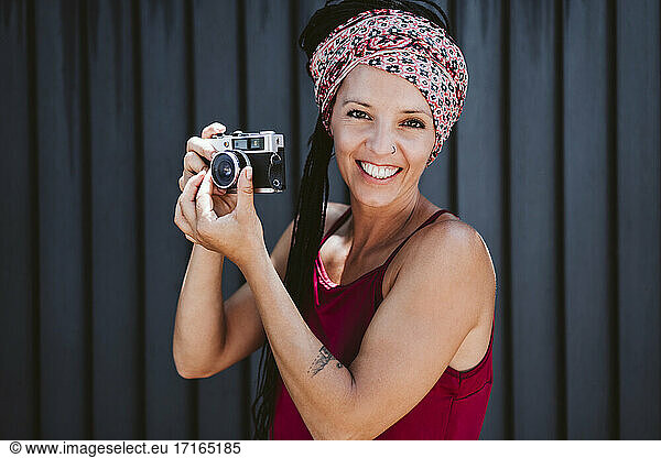 Smiling woman adjusting camera lens while standing against wall