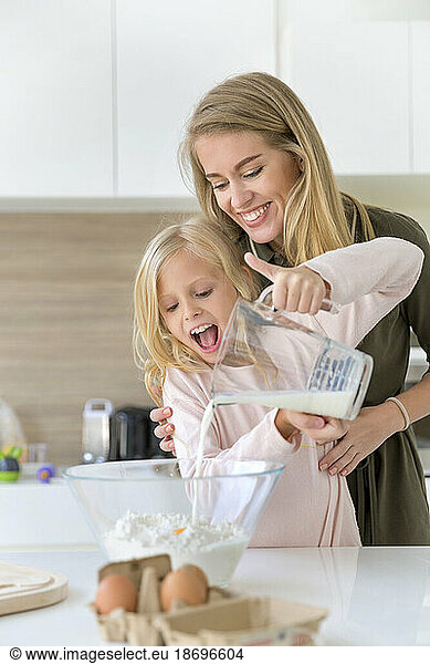 Smiling woman assisting daughter pouring milk in flour at home