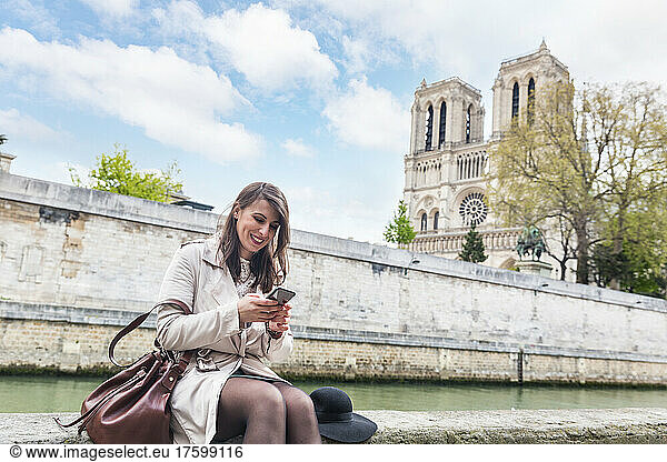 Smiling woman at riverbank using smart phone with Notre Dame Cathedral in background