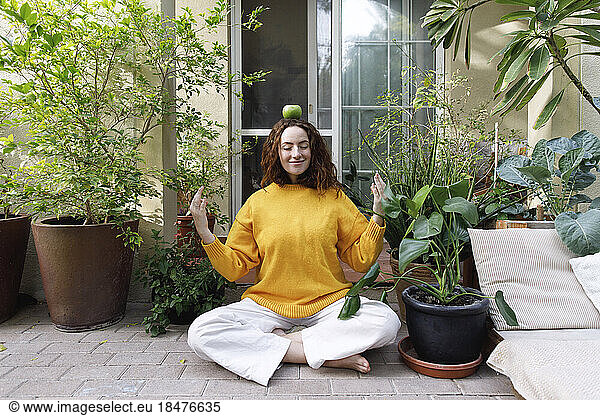 Smiling woman balancing green apple and meditating in backyard