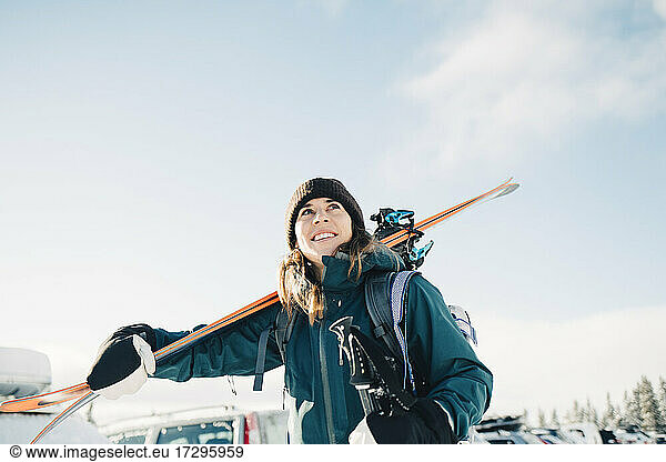 Smiling woman carrying skis during winter against sky