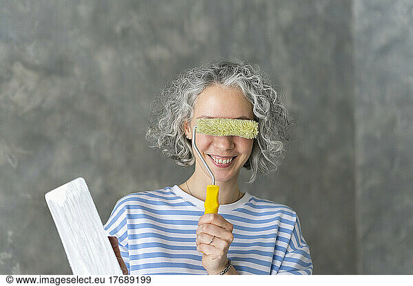 Smiling woman covering eyes with paint roller in front of wall