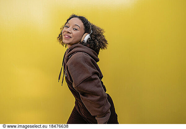 Smiling woman dancing with headphones by yellow wall