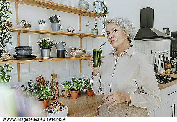 Smiling woman drinking detox juice in kitchen at home