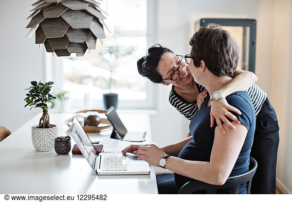 Smiling woman embracing girlfriend using laptop at table