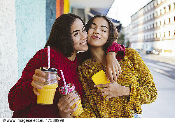 Smiling woman embracing twin sister with smart phone in city