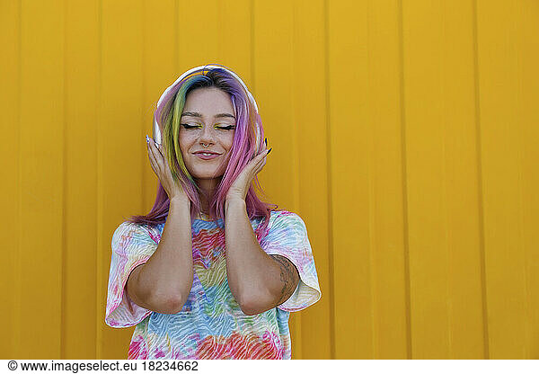 Smiling woman enjoying music listening through wireless headphones in front of yellow wall