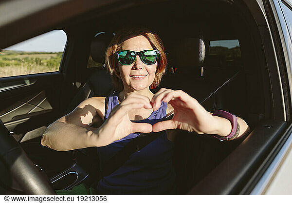 Smiling woman gesturing heart shape and sitting in car