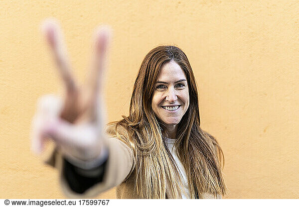 Smiling woman gesturing peace sign in front of wall
