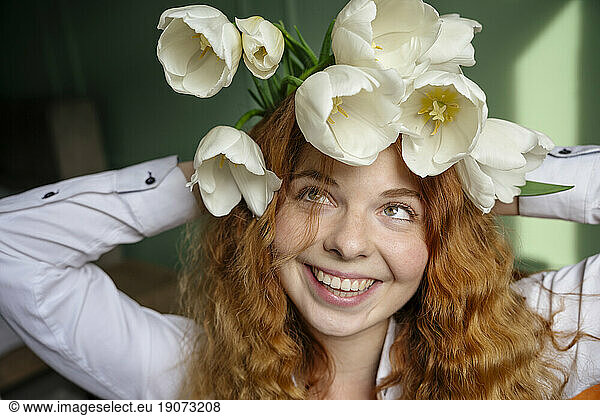 Smiling woman holding bunch of tulip flowers on head at home