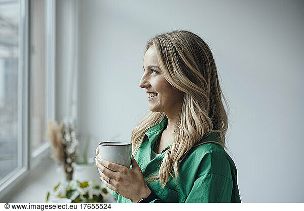 Smiling woman holding coffee cup looking out through window at home