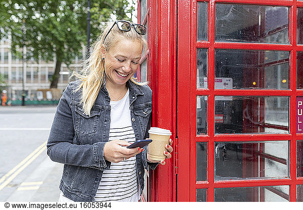 Smiling woman holding coffee cup using smart phone while standing by telephone booth