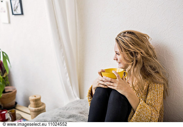 Smiling woman holding coffee cup while sitting on bed against wall