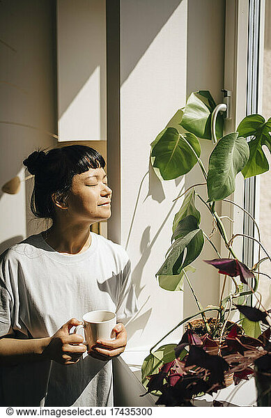 Smiling woman holding coffee cup while standing with eyes closed in sunlight at home
