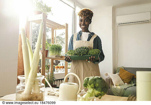 Smiling woman holding container of microgreens at home