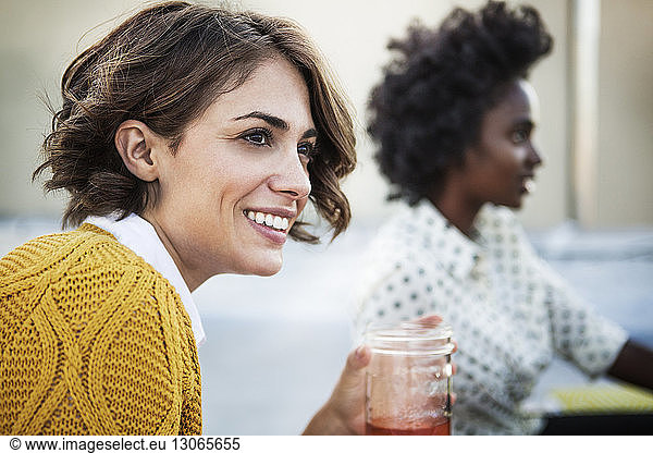 Smiling woman holding drinking glass with friend sitting in background
