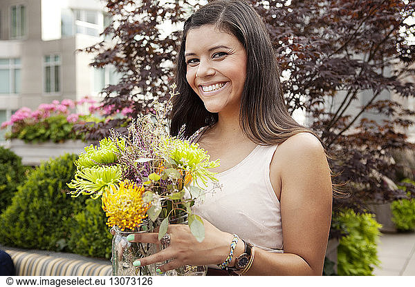 Smiling woman holding flowers jar while standing against plants
