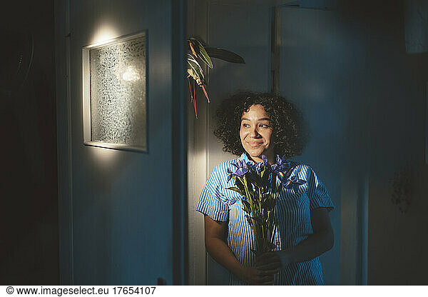 Smiling woman holding flowers leaning on wall at home