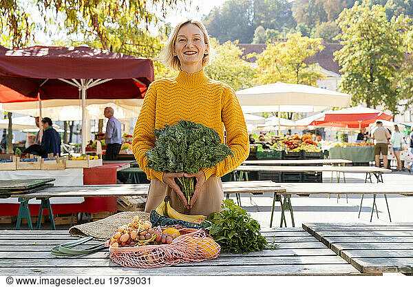 Smiling woman holding green leafy vegetables standing near table at farmer's market